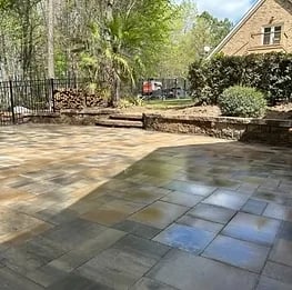 A brick courtyard with varied stone pavers in shades of gray and blue, featuring greenery and trees, adjacent to a residential building