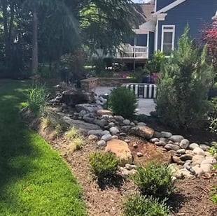 Landscaped residential backyard with rock garden bed, decorative boulders, green plants, and house with deck in background