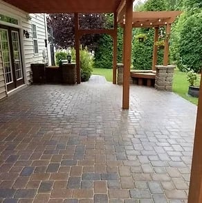 Backyard patio with brick pavers, wooden pergola, bench seating, and green lawn surrounded by landscaping