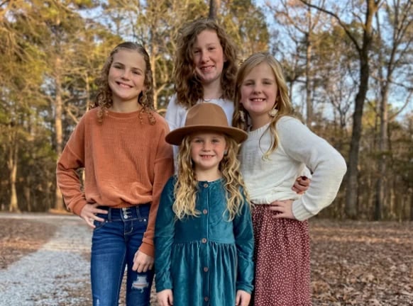 Four young girls smiling at the camera outdoors on a tree-lined path during autumn